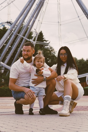 A family of three is posing together outdoors in front of a large Ferris wheel. The father, wearing a white shirt and denim shorts, crouches beside the mother, who is dressed in white and wearing glasses. Their young child stands between them pointing forward, smiling slightly. The setting appears to be a park or amusement area, with trees in the background.