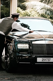 A person wearing a uniform and mask is polishing the hood of a luxury black car parked outdoors, with greenery visible in the background.