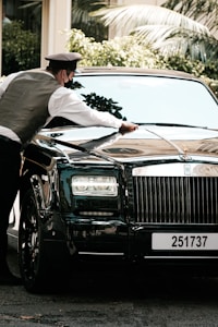A person wearing a uniform and mask is polishing the hood of a luxury black car parked outdoors, with greenery visible in the background.