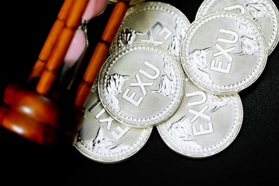 A group of silver coins inscribed with 'EXU' are scattered on a dark surface. An hourglass with wooden supports is partially visible, positioned diagonally on the left side.