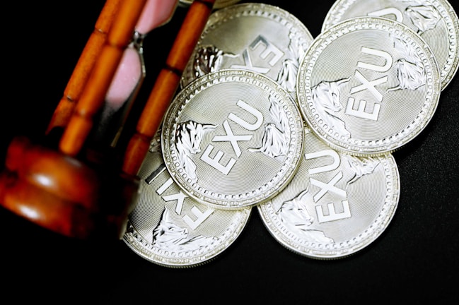 A group of silver coins inscribed with 'EXU' are scattered on a dark surface. An hourglass with wooden supports is partially visible, positioned diagonally on the left side.