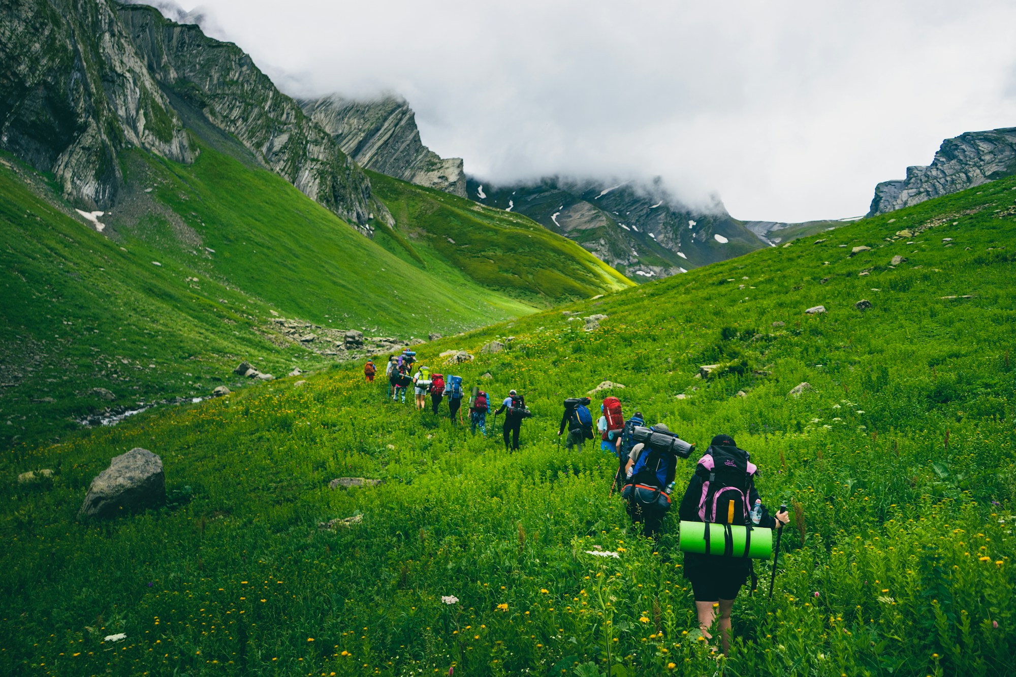 Small group of hikers with backpacks walking single file through a green alpine valley below cloud-covered ridges