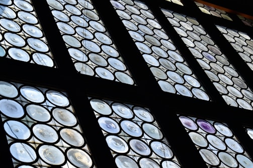 A set of round paneled windows featuring a pattern of circular glass pieces arranged in a grid. The glass has varying levels of transparency, with some displaying a slight iridescent effect. The framework is dark, contrasting with the light filtering through the windows.