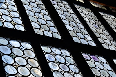 A set of round paneled windows featuring a pattern of circular glass pieces arranged in a grid. The glass has varying levels of transparency, with some displaying a slight iridescent effect. The framework is dark, contrasting with the light filtering through the windows.