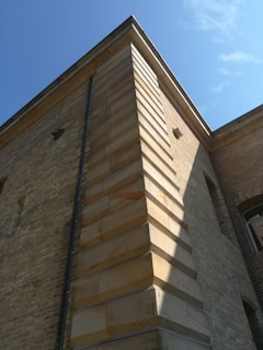A close-up view of a brick building corner with detailed masonry. The structure features alternating light and darker brick patterns. The perspective emphasizes the vertical lines and angles, while shadows create depth. The sky is clear and blue, suggesting a sunny day.
