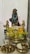 A religious altar setup featuring a blue deity statue adorned with a pearl necklace and a crown. Two smaller gold statues flank the main idol, and they are surrounded by offerings such as bananas, a mango, and a lime. There's a decoration of yellow flowers, and traditional brass utensils including lamps and a bowl with grains and coins.