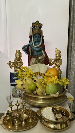 A religious altar setup featuring a blue deity statue adorned with a pearl necklace and a crown. Two smaller gold statues flank the main idol, and they are surrounded by offerings such as bananas, a mango, and a lime. There's a decoration of yellow flowers, and traditional brass utensils including lamps and a bowl with grains and coins.