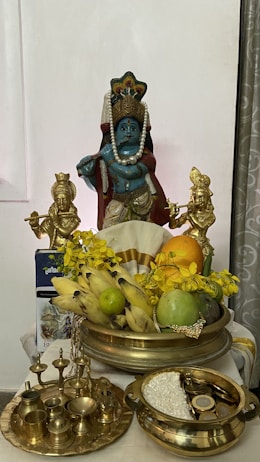 A religious altar setup featuring a blue deity statue adorned with a pearl necklace and a crown. Two smaller gold statues flank the main idol, and they are surrounded by offerings such as bananas, a mango, and a lime. There's a decoration of yellow flowers, and traditional brass utensils including lamps and a bowl with grains and coins.
