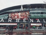 A large sports stadium with modern architecture is adorned with an iconic red and white logo. Large signage spells out 'ARSENAL' on the front, with an illuminated billboard featuring football players in red jerseys. People holding umbrellas gather around the stadium entrance, likely due to the rainy weather.