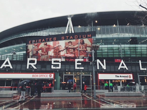 A large sports stadium with modern architecture is adorned with an iconic red and white logo. Large signage spells out 'ARSENAL' on the front, with an illuminated billboard featuring football players in red jerseys. People holding umbrellas gather around the stadium entrance, likely due to the rainy weather.