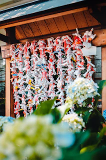 Numerous strips of paper are tied with red string and attached to a wooden structure, possibly part of a traditional Japanese wishing practice. In the foreground, blue and white hydrangeas add a touch of nature.
