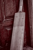 A wooden cricket bat leaning against an aged, textured wooden door. The bat has a light wooden color and visible grain patterns, with a taped handle. The background features a rich, dark wood with intricate panels and a slightly weathered appearance.