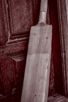 A wooden cricket bat leaning against an aged, textured wooden door. The bat has a light wooden color and visible grain patterns, with a taped handle. The background features a rich, dark wood with intricate panels and a slightly weathered appearance.