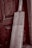 A wooden cricket bat leaning against an aged, textured wooden door. The bat has a light wooden color and visible grain patterns, with a taped handle. The background features a rich, dark wood with intricate panels and a slightly weathered appearance.