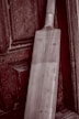 A wooden cricket bat leaning against an aged, textured wooden door. The bat has a light wooden color and visible grain patterns, with a taped handle. The background features a rich, dark wood with intricate panels and a slightly weathered appearance.