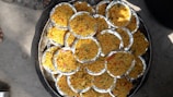 Multiple round foil plates filled with a dish primarily composed of yellow rice, interspersed with various small colorful ingredients such as red and green vegetables, are arranged aesthetically on a large circular tray. The scene is set outdoors, evident from the visible concrete ground and a shoe peeking into the frame.