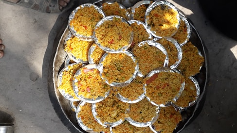 Multiple round foil plates filled with a dish primarily composed of yellow rice, interspersed with various small colorful ingredients such as red and green vegetables, are arranged aesthetically on a large circular tray. The scene is set outdoors, evident from the visible concrete ground and a shoe peeking into the frame.