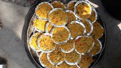 Multiple round foil plates filled with a dish primarily composed of yellow rice, interspersed with various small colorful ingredients such as red and green vegetables, are arranged aesthetically on a large circular tray. The scene is set outdoors, evident from the visible concrete ground and a shoe peeking into the frame.
