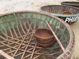 Large, round woven baskets, resembling boats, are placed on a sandy surface, featuring intricate wooden ribs and green mosaic-like detailing on the interiors. A smaller basket sits inside one of the larger baskets, and a long stick is positioned across it. The word 'PHUOC' is partially visible, painted in white on the exterior of one basket.