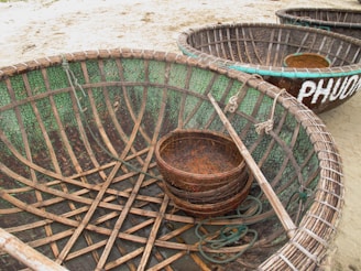 Large, round woven baskets, resembling boats, are placed on a sandy surface, featuring intricate wooden ribs and green mosaic-like detailing on the interiors. A smaller basket sits inside one of the larger baskets, and a long stick is positioned across it. The word 'PHUOC' is partially visible, painted in white on the exterior of one basket.