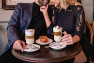 A couple is sitting closely together at a small round table, enjoying each other's company. Both are dressed in semi-formal attire. The table has two glasses of layered lattes, each topped with whipped cream and a sprinkle of cocoa powder, along with a croissant on a white plate in the center. They appear to be smiling and sharing a moment of affection.