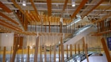 An interior space with modern architectural design featuring numerous suspended wooden beams and wooden accents. There are two escalators leading to different floors, and a few people can be seen walking around. The space is well-lit with natural and artificial lighting. Glass panels and large columns are part of the structural elements.
