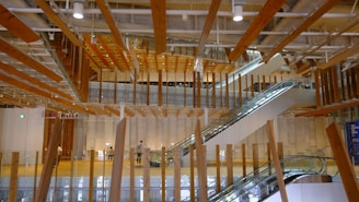 An interior space with modern architectural design featuring numerous suspended wooden beams and wooden accents. There are two escalators leading to different floors, and a few people can be seen walking around. The space is well-lit with natural and artificial lighting. Glass panels and large columns are part of the structural elements.