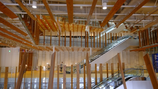 An interior space with modern architectural design featuring numerous suspended wooden beams and wooden accents. There are two escalators leading to different floors, and a few people can be seen walking around. The space is well-lit with natural and artificial lighting. Glass panels and large columns are part of the structural elements.