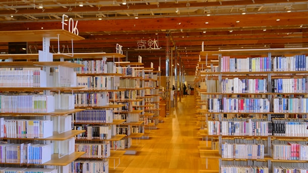 A spacious library interior with wooden bookshelves filled with a variety of books. The neatly organized shelves are placed symmetrically on a polished wooden floor, creating a sense of order and calm. Overhead, warm lighting illuminates the space.