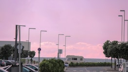 A seaside parking lot with several parked cars. A camper van is positioned near the waterfront, and modern, tall street lamps line the parking area. The sky is painted in shades of pink with soft clouds, suggesting a sunrise or sunset. Trees are arranged alongside the lot, adding natural greenery to the scene.