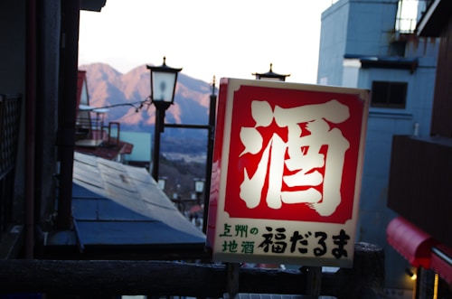 A Japanese sign with bold red and white characters prominently displayed in the foreground. In the background, there are traditional street lamps, buildings with sloped roofs, and a view of distant mountains under a clear sky.