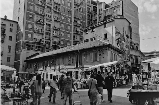 A busy street market scene with several people walking and browsing stalls that are set up with various items for sale. The area is surrounded by tall residential buildings with balconies and numerous windows. An old building with shuttered windows and a sloping roof stands at the forefront, and there is a mural on one side. The atmosphere is lively.