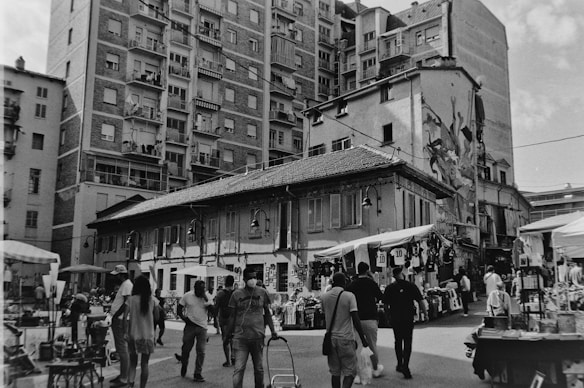 A busy street market scene with several people walking and browsing stalls that are set up with various items for sale. The area is surrounded by tall residential buildings with balconies and numerous windows. An old building with shuttered windows and a sloping roof stands at the forefront, and there is a mural on one side. The atmosphere is lively.