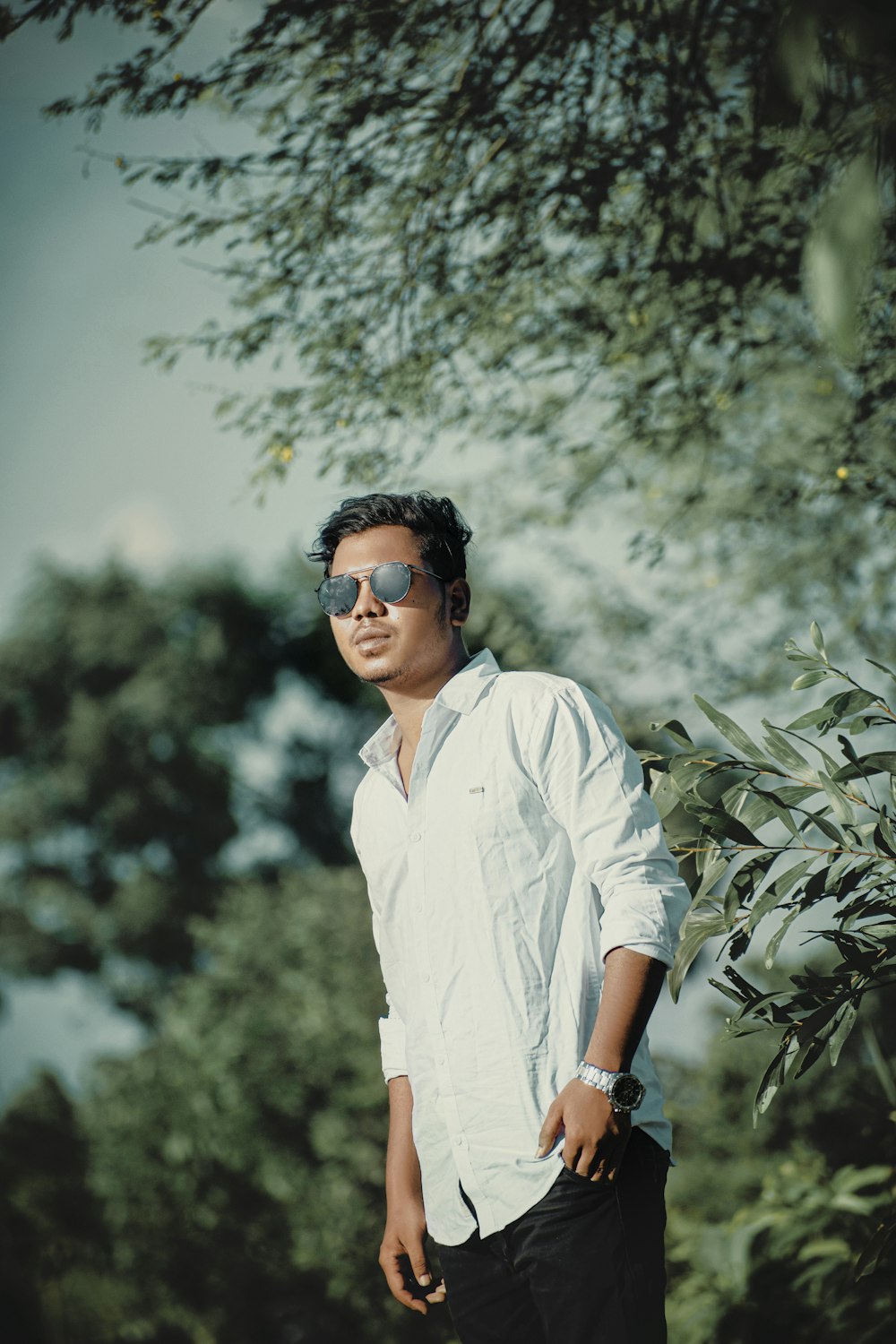 woman in white button up shirt wearing black sunglasses standing near green leaf tree during daytime