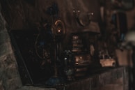 A collection of vintage and antique objects is displayed on a dimly lit stone mantel. There are metallic vases, lamps, and what appears to be an old toy car, with intricate designs and muted colors. The lighting is low, creating a mysterious and nostalgic atmosphere.