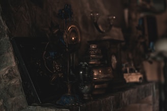 A collection of vintage and antique objects is displayed on a dimly lit stone mantel. There are metallic vases, lamps, and what appears to be an old toy car, with intricate designs and muted colors. The lighting is low, creating a mysterious and nostalgic atmosphere.