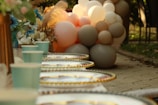 A festive outdoor setting with neatly arranged paper cups and golden-rimmed plates on a table. In the background, there is a cluster of pastel-colored balloons and a bouquet of flowers with blue and white petals. The scene has a warm and inviting atmosphere with soft natural lighting.
