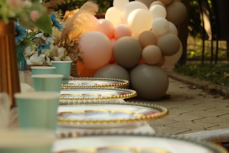 A festive outdoor setting with neatly arranged paper cups and golden-rimmed plates on a table. In the background, there is a cluster of pastel-colored balloons and a bouquet of flowers with blue and white petals. The scene has a warm and inviting atmosphere with soft natural lighting.