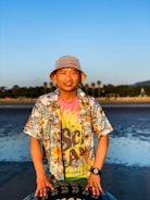 A person wearing a colorful floral shirt, a bright tie-dye t-shirt, and a beige bucket hat stands outdoors near the beach. The background features a calm body of water, palm trees, and a clear blue sky. The person appears to be smiling and has their hands resting on a patterned object in front of them.