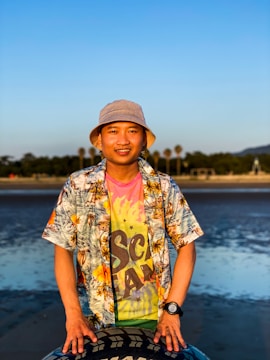 A person wearing a colorful floral shirt, a bright tie-dye t-shirt, and a beige bucket hat stands outdoors near the beach. The background features a calm body of water, palm trees, and a clear blue sky. The person appears to be smiling and has their hands resting on a patterned object in front of them.