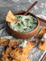 A wooden bowl filled with a creamy dip garnished with fresh parsley set on a wooden tray. Surrounding the bowl are several pieces of crispy pita chips, some topped with sesame seeds and herbs. One chip is placed inside the dip, partially covered.