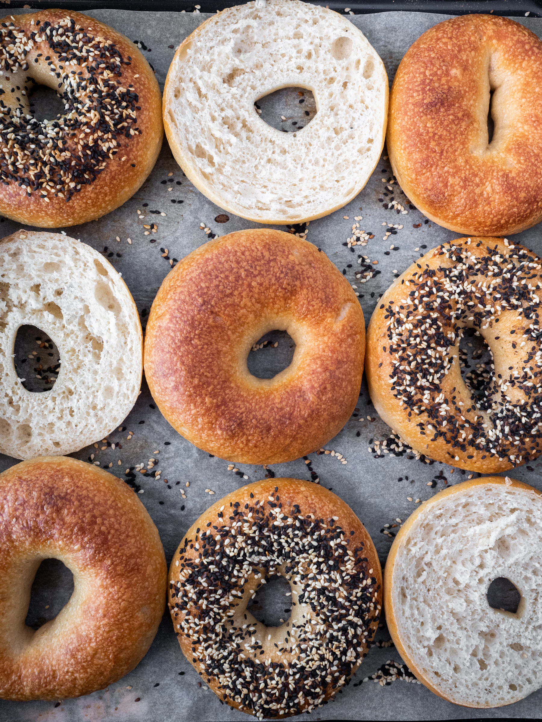 An overhead grid of plain and seeded bagels on parchment, some halved to show a dense, even crumb