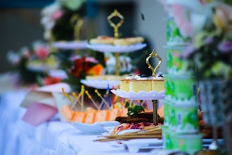 A beautifully arranged table with an assortment of cakes and desserts displayed on tiered stands. The stands are adorned with decorative handles, and there are various colorful treats, including pastries and fruit skewers, accompanied by floral arrangements in the background.