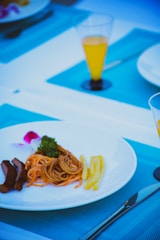 A white plate holds a small portion of pasta, a few strips of grilled meat, and a serving of fries, garnished with a sprig of parsley and a pink flower petal. The setup is on a blue tablecloth, and there are two glasses of orange juice visible.