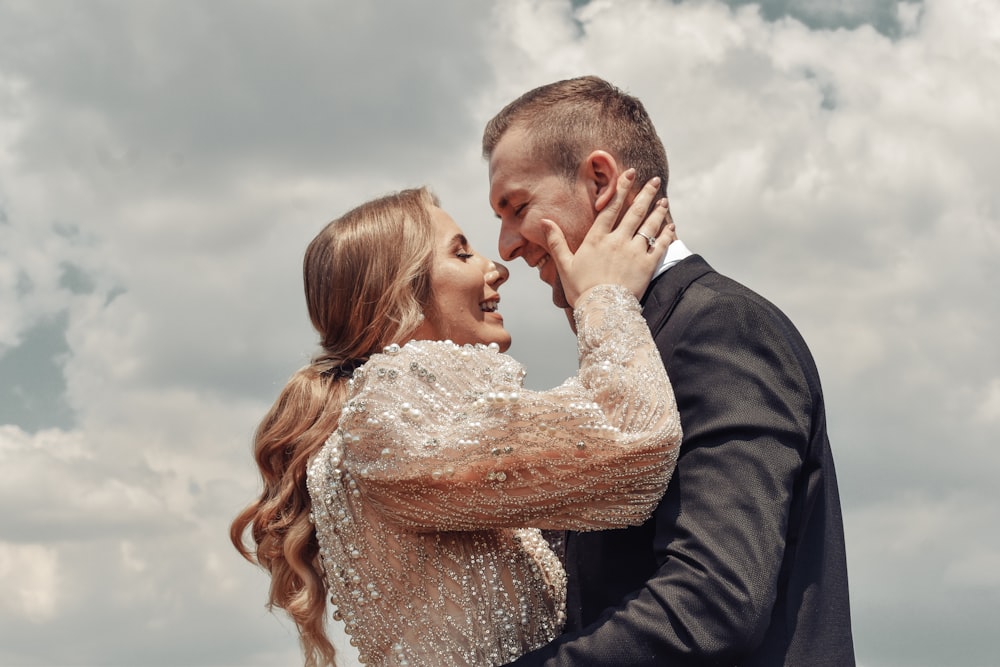 man in black jacket kissing woman in white floral lace dress under cloudy sky during daytime