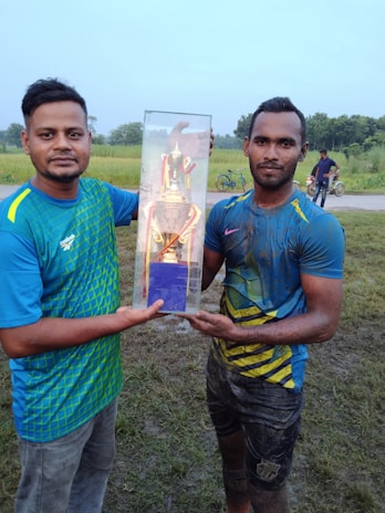 Two men are standing outdoors on a grassy area. They are holding a trophy encased in a transparent rectangular box. One man has a clean appearance while the other is covered in mud, suggesting participation in a sports event. In the background, a rural setting with greenery and a road is visible, along with people cycling and walking.