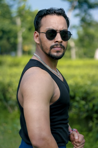 A man wearing a black tank top and dark sunglasses, standing outdoors with a confident pose. He has short dark hair and a beard, and is accessorized with a silver chain around his neck. The background features green foliage, suggesting a park or garden setting.
