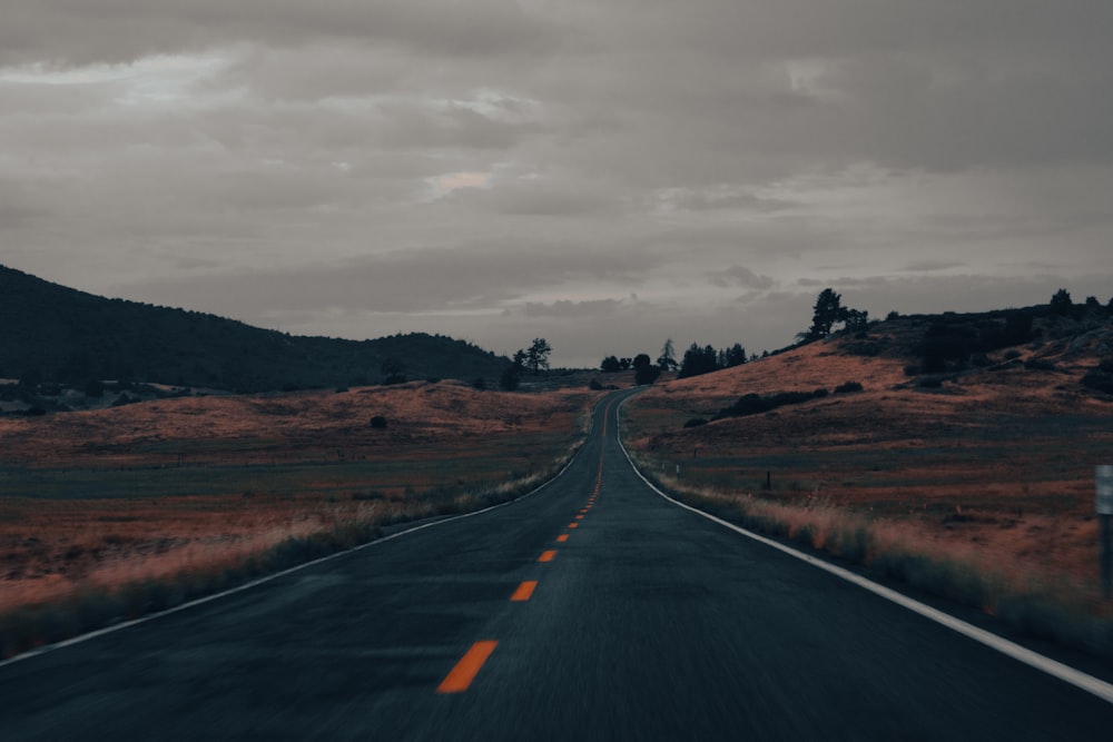 black asphalt road in between brown grass field under gray cloudy sky during daytime