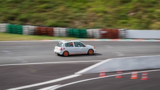 A small white car with orange rims is speeding around a curved racetrack. The track is bordered by stacks of multicolored tires and orange traffic cones are positioned along the inner edge. The car appears to be moving quickly, evident from the motion blur.