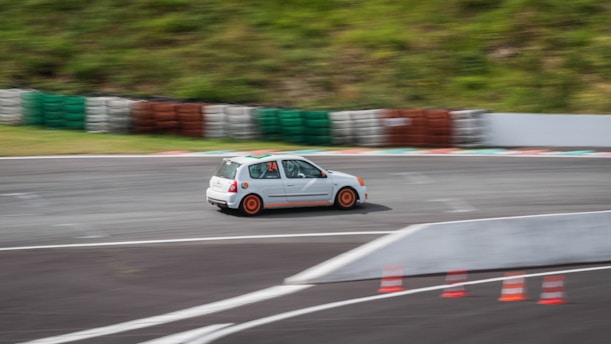 A small white car with orange rims is speeding around a curved racetrack. The track is bordered by stacks of multicolored tires and orange traffic cones are positioned along the inner edge. The car appears to be moving quickly, evident from the motion blur.